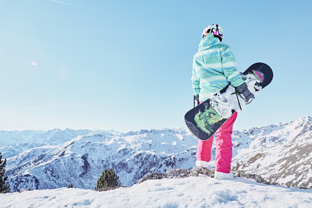 Back view of female snowboarder wearing colorful helmet, blue jacket, grey gloves and pink pants standing with snowboard in one hand and enjoying alpine mountain landscape - snowboarding concept
